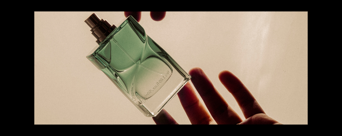 close up of a light blue perfume bottle suspended in mid-air being held by fingers on two hands against a cream background.