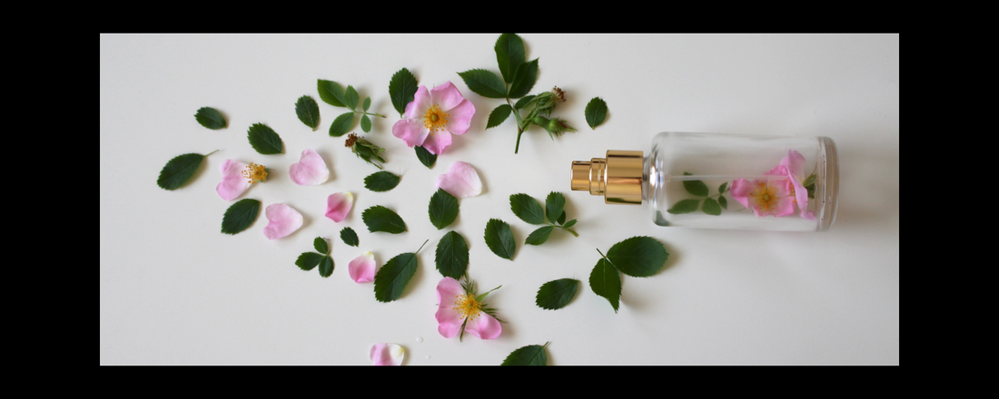 Small perfume bottle lay flat on a light grey table with flower petals and leaves spread across the table.